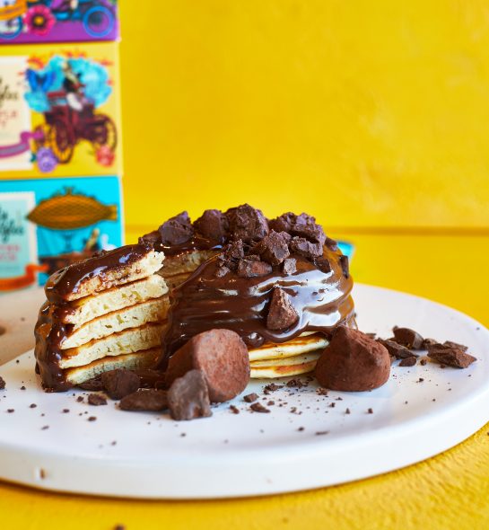 A stack of pancakes on a white plate,covered in melted chocolate sauce, on a yellow background, next to a box of Monty bojangles chocolate truffles.