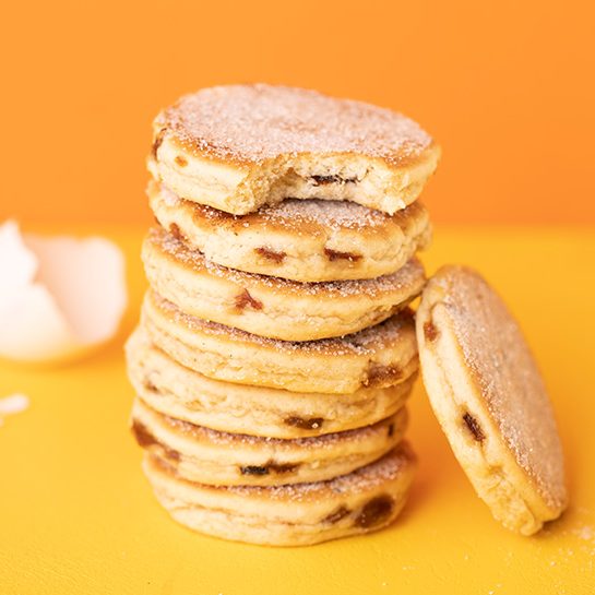 A stack of eight welsh cakes, coated in sugar, on a yellow background, with one extra cake resting to the side of the stack.