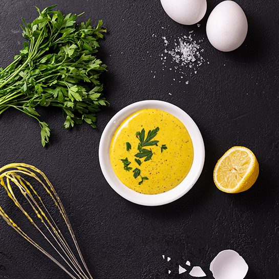 A white pot filled with vibrant yellow mayonnaise on a black background, surrounded by egg shells, a whisk and parsley leaves