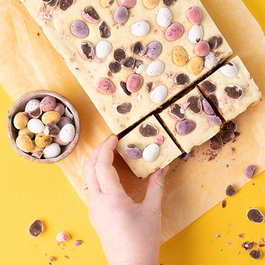 A hand reaches for a block of blondies, cut into slices, topped with white chocolate and mini eggs, against a yellow background