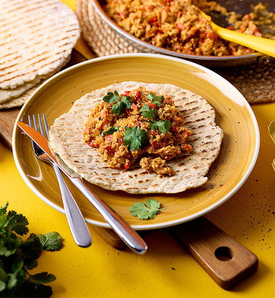 A pile of tomatoey Turkish eggs sit on a flatbread on a round yellow plate, which all sits on a wooden chopping board. The eggs are topped with a handful of green coriander.