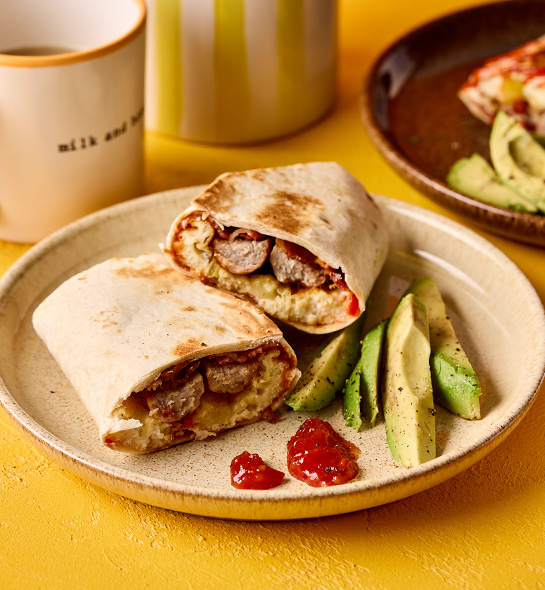 Breakfast burritos cut in half on a plate so that the egg, sausage and bacon on the inside is showing. The wholemeal wrap is toasted, with a dollop of ketchup and some sliced avocado sitting to the side.