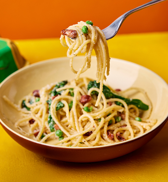 A bowl of spaghetti carbonara mixed with peas and broccoli, on a yellow table.