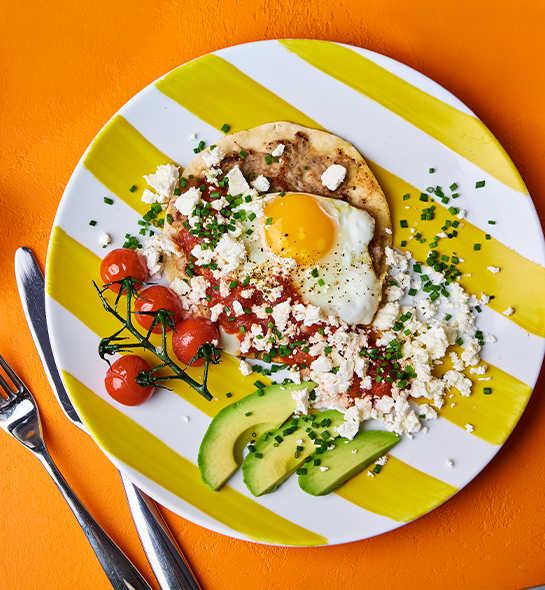Smashed sausage taco on a yellow and white stripy plate, topped with a fried egg, vine tomatoes, avocado slices and crumbled feta.